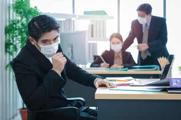 Flu sick businessman in suit wearing face mask and coughing while working in office, colleague wearing mask too for protecting  themselves against virus, decreased risk and preventing the spread of co