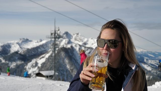 Medium closeup of a Caucasian woman with brown hair wearing sunglasses and drinking beer at a skiing resort with skiing cable cars and snow covered mountains in the background
