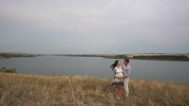 A happy man gently strokes the tummy of a pregnant wife standing in a field against the background of a large lake in windy weather.