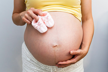 Isolated portrait of a pregnant woman holding pink crochet baby shoes and showing her baby bump over white background