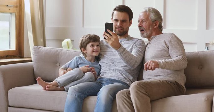 Smiling young man sitting on sofa with little child son and middle aged pleasant father, showing funny mobile application. Happy three male generations family posing for selfie shot on smartphone.
