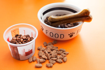 Measuring cup, bowl with dry food and toys for dogs on the orange background. Concept pet care, playing and training. Top view. Space for a text.