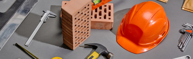 panoramic shot of bricks, hammer, angle keys, orange helmet, calipers and putty knife on grey...