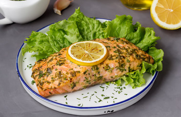 One piece of salmon fillet baked with lemon and herbs. Dark background, white plate.