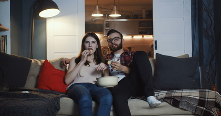 Naklejka premium Happy young casual European romantic couple watching TV series at home, smiling and talking, eating popcorn slow motion