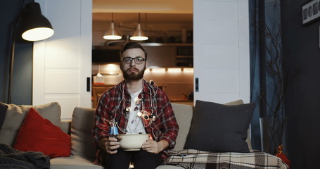 Young Caucasian man is sad and depressed watching movie at home, eating snacks alone at empty house...