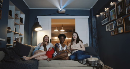 Cheerful smiling multiethnic female friends watch film together at home, laugh and talk eating...