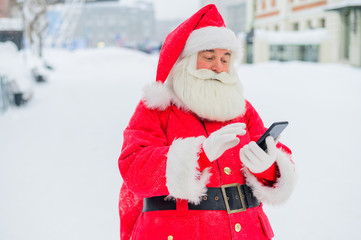 Fototapeta na wymiar Portrait of a funny funky fat modern Santa Claus use his mobile phone. An elderly man in a suit before Christmas communicates on a smartphone.