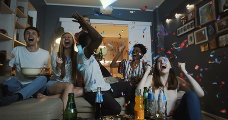 Happy young multiethnic fans raise hands and shout watching sports game at home on TV with...