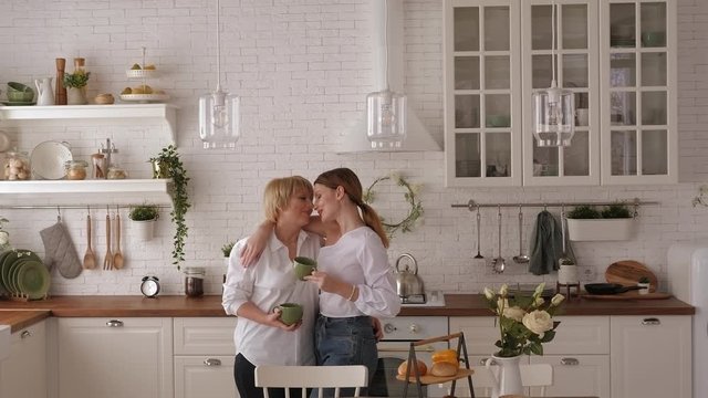 Portrait of an adult mother and daughter hugging in the kitchen, they drink tea and talk. A happy elderly mother embraces her daughter in the bright kitchen, they hold green mugs of tea in their hands