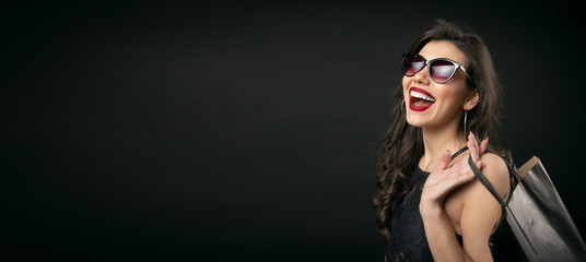 Overjoyed young woman in sunglasses holding shopping bags on shoulder isolated over black background