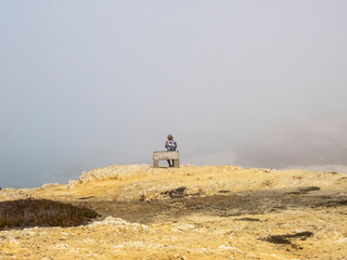 Una mujer sentada en un banco de piedra en un acantilado de la costa portuguesa en un día de niebla