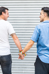 Two men with lgtb flag bracelets holding hands while looking at each other in front of a metal blind