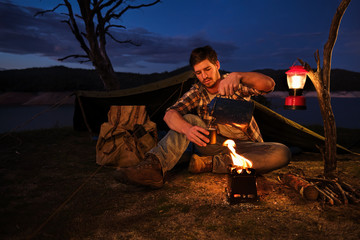 Man in hiking boots next to a camp fire pouring coffee 