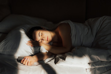 Sunny morning at home. Tender woman lying on bed sleeping on side calm. Horizontal shot of relaxed carefree female under white bedclothes in bed at bedroom, sees pleasant dreams, keeps eyes close. 