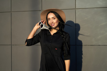 Autumn trendy collection. Happy beautiful brunette girl wearing stylish grey dress holding fashionable hat in the hands. Model in trendy outfit posing on grey wall background. Outdoor portrait.