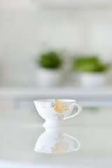 Working alone at the office, coffee cup on a white table over blurred clean kitchen with no one and no clients in a cafe, a reflection of the floral cup is reflecting from the table