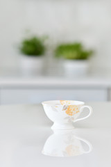 Working alone at the office, coffee cup on a white table over blurred clean kitchen with no one and no clients in a cafe, a reflection of the floral cup is reflecting from the table