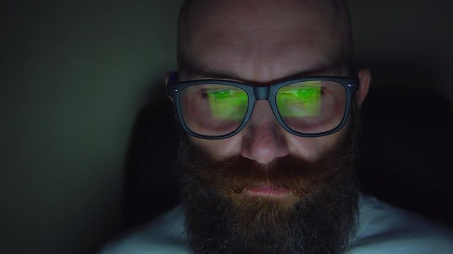 Portrait of bearded man in eyeglasses browsing website at late night, reflection at glasses