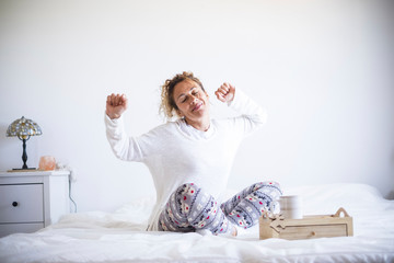 Fototapeta na wymiar Early morning wake up concept with happy cheerful caucasian beautiful adult woman stretching and smiling - white colors and light in the bedroom athome or hotel - stay home quarantine