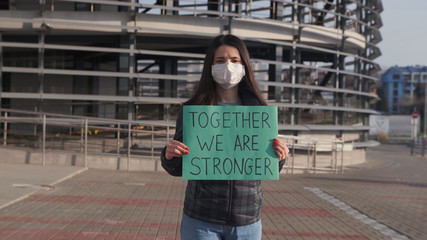 A young woman in a protective mask stands on the street and holds a placard with the inscription together We are stronger. A call for solidarity.