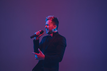 Young caucasian musician, performer singing on gradient purple-blue background in neon light. Concept of music, hobby, festival. Joyful party host, DJ, stand upper, singer. Colorful portrait of artist