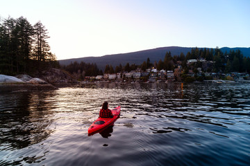 Adventurous Girl Paddling on a Bright Red Kayak in calm ocean water during a vibrant and colorful sunset. Taken in Indian Arm, Deep Cove, North Vancouver, British Columbia, Canada.