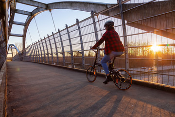 Caucasian Woman Riding a Bicycle on a Pedestrian Bridge over the Highway during a sunny sunset. Taken in Surrey, Vancouver, British Columbia, Canada.