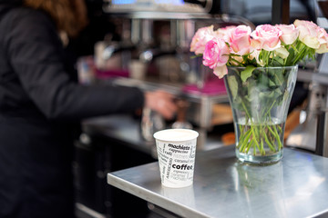 Paper cup with cappuccino coffee in right bouquet of pink-white roses in glass vase with water and coffee maker on background