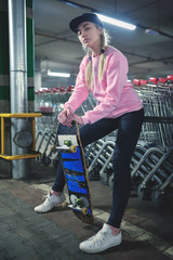 Young female model posing with skate board. Shopping carts - supermarket underground car park. Street style fashion - black cap and pink blouse. Youth lifestyle.