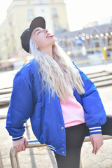 Happy, smiling woman sitting on railing and having fun. Trendy hairstyle - white blonde hair. Youth style fashion - black cap, blue baseball jacket.