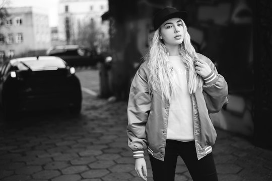 Black and white photo blonde woman, posing on the street. Youth style fashion - black cap and baseball jacket. Town background - buildings, cars on the street.