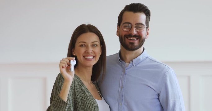 Close up head shot overjoyed young family couple looking at camera demonstrating keys from new flat. Happy 30 wife embracing husband, feeling excited about moving in apartment, real estate concept.