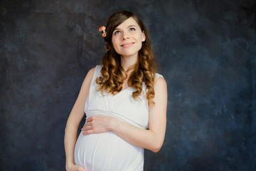 Fototapeta na wymiar Portrait of dreamy pregnant young woman in white dress. Maternity concept. Attractive caucasian expectant mom touching her belly, dark wall on background.