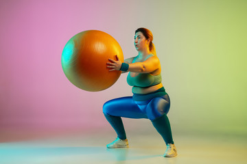 Young caucasian plus size female model's training on gradient purple green background in neon light. Doing workout exercises with fitball. Concept of sport, healthy lifestyle, body positive, equality.