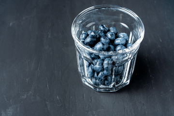 Beautiful tasty raw blueberry with a transparent glass cup on a dark black concrete background.