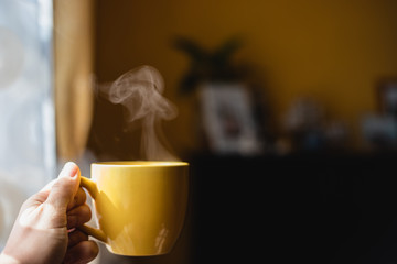 Yellow cup with hot streaming tea drink in hand near window with home interior background in morning light