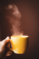 Yellow cup with hot streaming tea drink in hand with dark pink background in beautiful morning light