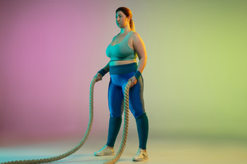 Young caucasian plus size female model's training on gradient purple green background in neon light. Doing workout exercises with ropes. Concept of sport, healthy lifestyle, body positive, equality.