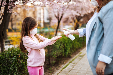 Obraz premium Mother and daughter standing in park, wearing protective masks and disinfecting hands with alcohol.