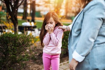 Cute little girl sneezing in park while she is walking in park with her mother. Allergy, flu, virus concept..