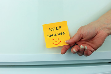 A hand holding a sticky note on a fridge door with the words keep smiling.