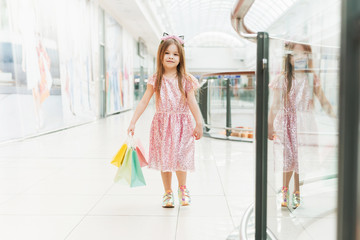 Portrait of a little happy girl in the mall. A smiling laughing girl in a pink dress with a cute rim with ears and with multi-colored bags in her hands is walking around the mall