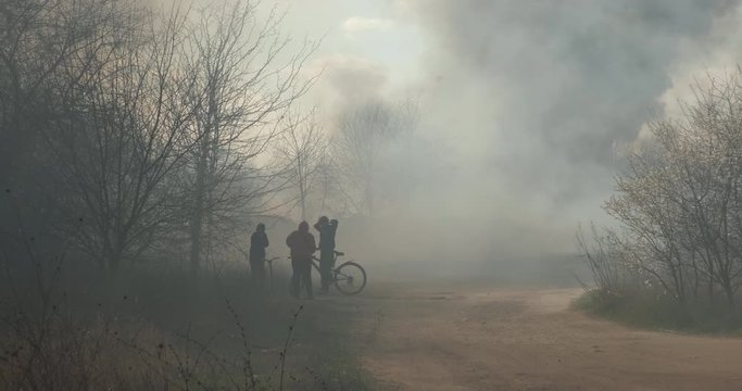 Clouds smoke fire in city park on blue sky, bright sun disk and sun rays, arson on background of trees in spring. Teenagers in park are looking at smoke. Protest, criminal incident. Effects. Flames