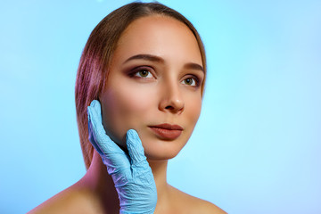 Doctor's hand in glove touch the face of young woman isolated on blue background. Healthcare, cosmetology, medical and plastic surgery concept