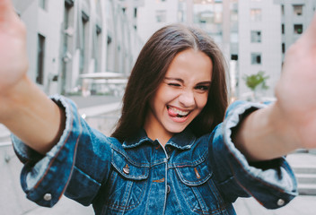 Young positive stylish hipster girl winking and showing tongue while making selfie on camera....