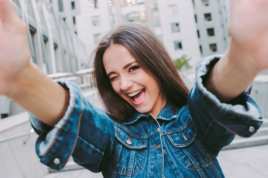 Young happy stylish woman winking and smiling while taking selfie on smart phone. Beautiful stylish cheerful hipster girl making portrait photo on camera. Having fun and making pictures on city street