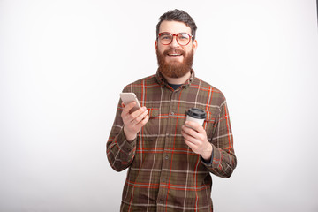 Fototapeta premium Portrait of cheerful young man with beard holding smartphone and cup of coffee to go
