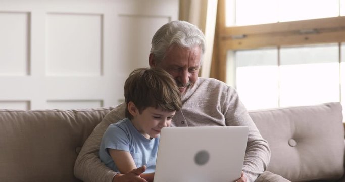 Happy senior mature grey haired man embracing small kid, using computer together. Smiling little child son watching funny movie cartoons with affectionate middle aged grandfather on laptop at home.