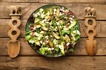 Green salad with avocado and grilled beans and courgettes on a green plate set against a wooden table.  Wooden salad servers with elephant carved handles placed on either side of the salad plate.